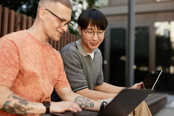 Candid shot of smiling Asian male IT programmer working on laptop collaboratively with colleague...