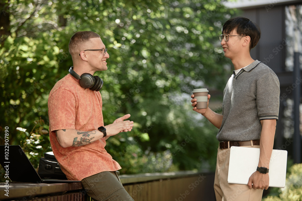 Poster side view of smiling it developer with headphones chatting with cheerful partner holding laptop and 