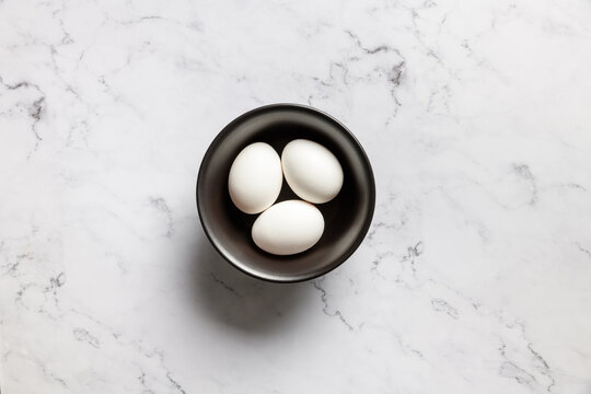 Top view of three white eggs in a vintage black bowl on a marble background.