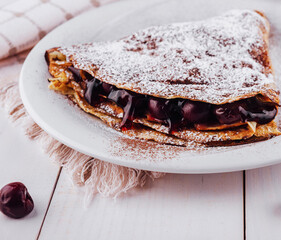 Delicious homemade crepes filled with cherry jam and cocoa powder on white rustic wooden background