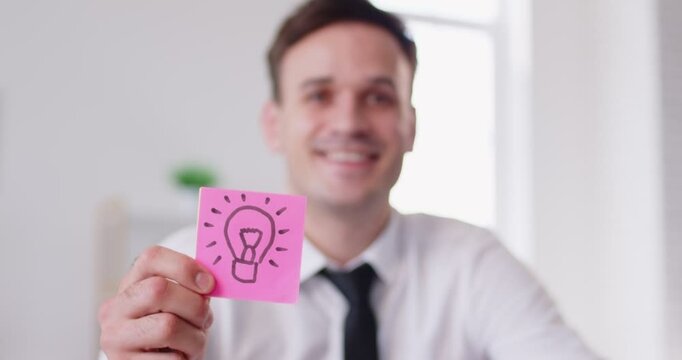 Happy businessman pointing upward with finger, illustrating a eureka moment as he shows a light bulb lamp drawn on a sticky note. Creativity, inspiration, and the spark of a new idea.