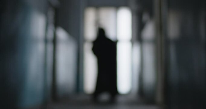 Blurred defocused silhouette of person in grim reaper death costume in black cloak holding scythe walking indoors on dark background. Halloween and horror concept. 4k video. Slow motion video.