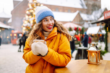 Happy woman drinks a hot drink at a Christmas market decorated with festive lights. Festive Christmas fair, winter holidays concept.