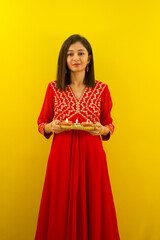 Happy young Indian woman in traditional dress holding a plate of oil lamps on Diwali festival, looking at camera on yellow background.