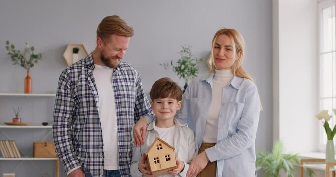 Family ownership. Happy family consisting of father, mother, and son proudly show off keys to their new home. Joyful family of three holding house model and keys and smiling at camera.