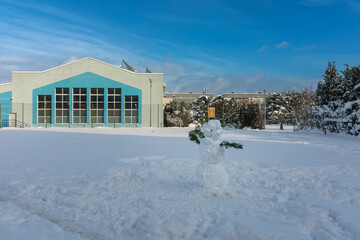 cute happy snowman on snow-filled football field. day during winter. school and street in the background.