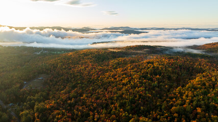 Aerial view of a mountain range covered in vibrant fall foliage, with mist and fog drifting over the colorful forested hills, capturing the beauty of the autumn season.