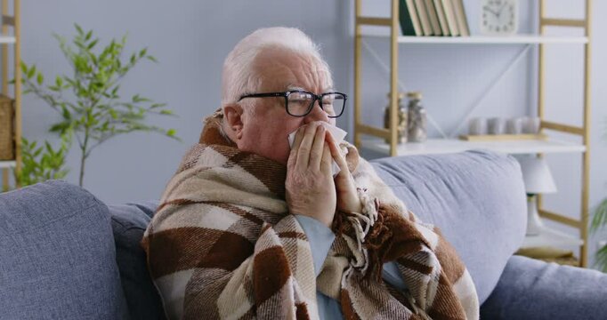 Senior man, feeling unwell, is wrapped in a blanket, sitting on a sofa at home. He is coughing or sneezing into a tissue, possibly experiencing symptoms of a cold, flu, or respiratory virus.