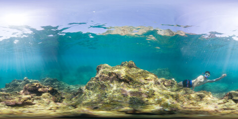 A person snorkeling near a rocky coral formation underwater, captured in a 360-degree view. The sunlight penetrates the clear water, highlighting the rocky terrain and marine life.