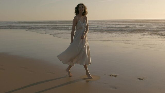 A woman is walking barefoot on a beach during sunset, wearing a flowing, backless dress. She is gracefully holding one hand up to her hair, creating a serene and relaxed atmosphere. 