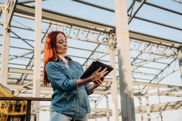 Holding notepad. Young redhead woman is standing near unfinished building, construction