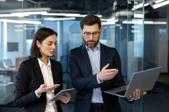Colleagues in formal attire engage in business discussion using laptop and tablet. Creative workspace setting highlights team communication and project management in modern office.