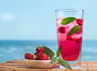 Raspberry lemonade with mint and ice on a wooden table with the sea on the background.