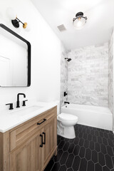 A bathroom with a white oak cabinet, black and gold light fixtures, black marble picket tile flooring, and a marble subway tile shower. No brands or labels.