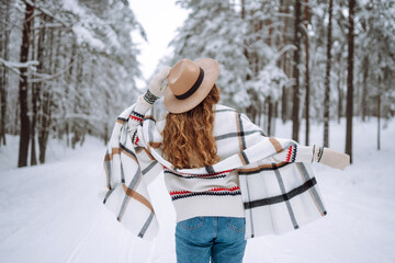 Back view of woman standing among snowy trees in winter forest and enjoying first snow. Wearing hat, plaid scarf and coat. Wanderlust and boho style