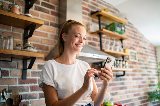 Smiling young woman using smartphone in modern kitchen