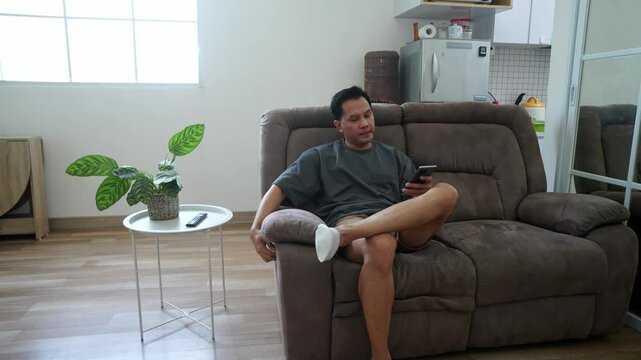 Young Asian Man Yawning And Sleeping On Reclining Sofa In Front Of A TV At Home