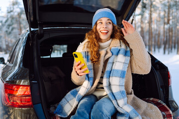 Young Woman sitting in the trunk of her and using phone in winter time. Concept of technology,...