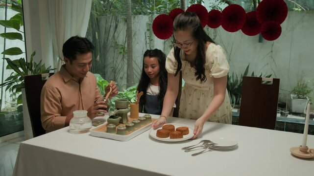 Medium long shot of Asian family of three celebrating mid-autumn with traditional mooncakes and herbal tea at home