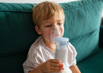 Cute little boy sitting and holding a nebulizer mask leaning against his face, respiratory tract...