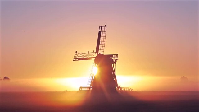 High quality 4K film of traditional Dutch wind mill and wind turbine at orange and pink fog sunrise with sun rays moving behind mill