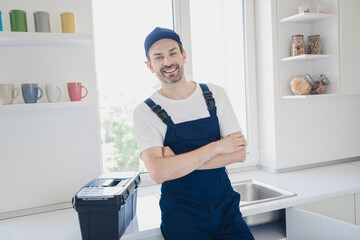 Smiling handyman in blue overalls standing confidently in bright kitchen with toolbox ready for...