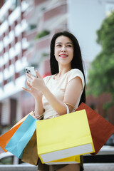 A cheerful young Asian woman, stylishly dressed, smiles excitedly while holding shopping bags....