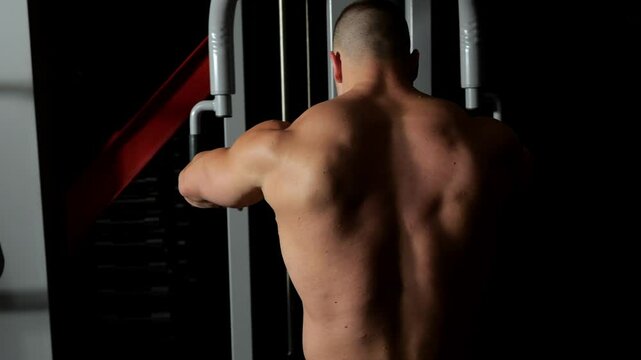 A bodybuilder uses the fly simulator to train his pectoral muscles. Upper body workout in a dark gym, view from the back. A handsome bodybuilder trains in the gym on the fly press simulator.