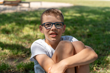 Smiling boy wearing glasses sits on the grass in a park on a sunny day, enjoying the outdoors and relaxation. High quality photo