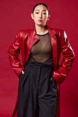 Fototapeta premium Fashion asian woman in red leather jacket and black pants posing with hands on hips against red background