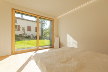 View of a bedroom with cardboard home staging furniture, a very bright window overlooking nature, green lawn and trees. No one inside.
