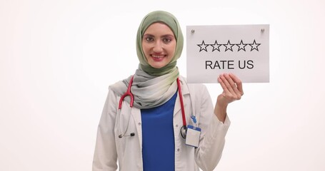 Happy woman doctor shows Rate Us sign pointing with hand against white. Muslim female medic in hijab encourages patients to leave positive review