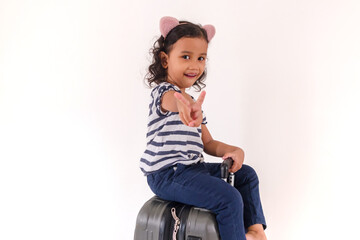 A smiling little Asian girl sitting on a suitcase and showing two fingers or victory sign, on a white background