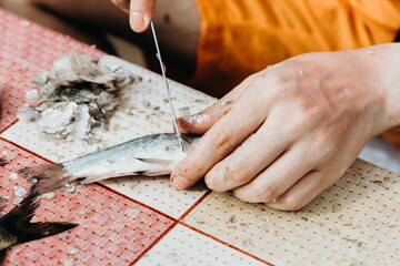 A young man cleans fish from scales while sitting at a table.