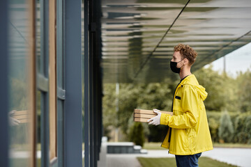 A delivery person in a black mask and yellow jacket arrives at an office building with pizza boxes...