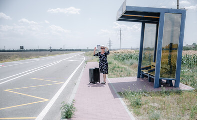 An elderly woman with a suitcase stands at the bus stop.