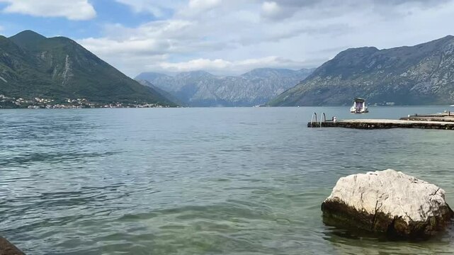 Time-lapse of Prčanj from Radimiri with distant SUP paddler and motorboats in Kotor Bay, framed by scenic mountains and calm waters, capturing serene natural beauty in light clouds and clear skies