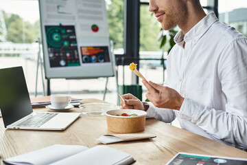 A redheaded man in a white shirt engages with his project while savoring a delicious meal at a stylish desk.