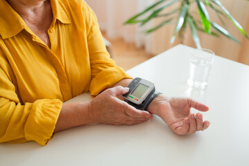 Healthy woman self checking blood pressure using machine. Senior woman with hypertension measuring blood pressure herself at home. Heart rate measurement. Glass of water on the table