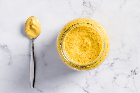 Top view of a spoon and open jar full with musterd on a marble background.