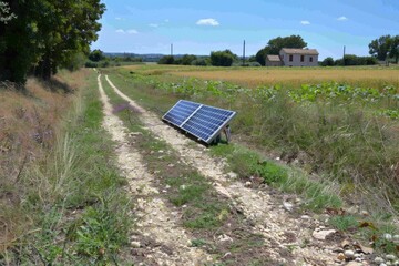 A pair of solar panels sit on a dirt road in a rural setting, surrounded by lush greenery and a distant farmhouse.