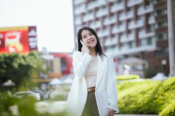 Fototapeta premium Active Asian woman in suit standing outdoors in city holding smartphone and laptop. She represents the future of finance and investment, keeping updated on news and opportunities.