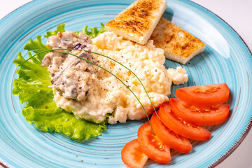 Scrambled eggs with mushrooms and crispy toast, garnished with lettuce and tomatoes, on a turquoise plate, on a white table with a burgundy napkin and cutlery. Delicious and healthy breakfast.