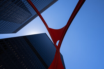 Looking up from below, sleek modern skyscrapers are framed by a bold, red steel abstract structure against a clear blue sky. The contrasting elements create a striking, upward perspective.