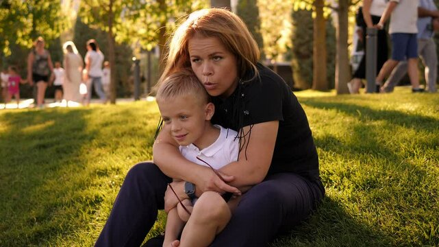 A happy mother and son are sitting on the green grass in the city park and hugging, a lot of people are walking in the background.