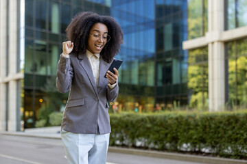 Fototapeta premium Confident African American businesswoman celebrates success while reading exciting news on phone. Standing outside modern office building, she displays joy and victory.
