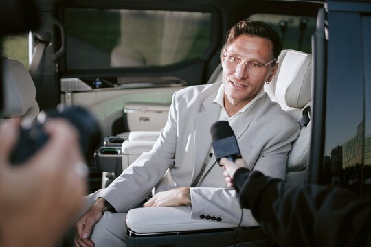 Medium shot of male celebrity in glasses speaking with TV media giving interview in car while journalists taking exclusive comments of superstar, shot in cool tone, copy space
