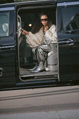 Glamorous Middle Eastern female celebrity sitting in car wearing trendy silvercore pants and boots...