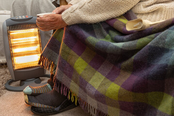 Elderly pensioner trying to keep warm near an electric heater with blanket and a warm drink. In...
