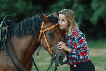 Fototapeta premium Happy blonde with horse in forest. Woman and a horse walking through the field during the day. Dressed in a plaid shirt and black leggings.